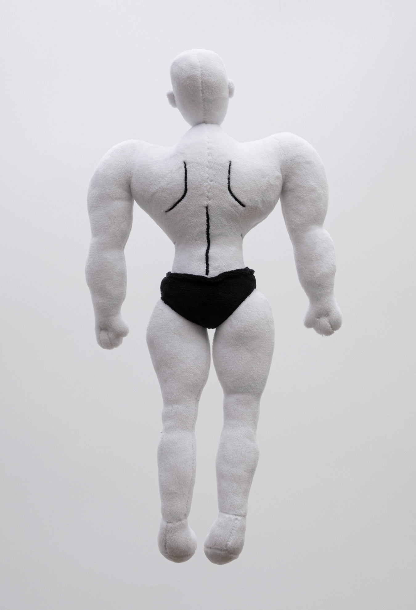 The back of a plush figure of a muscular male character with white skin, bald head and black briefs against a white background.